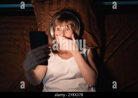 Worried mature woman watching bad online content in a smartphone lying on a bed at night in her bedroom. Top view Stock Photo