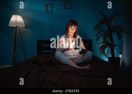 Sleepy exhausted lady lying in bed using smartphone. Sad woman using mobile phone late at night in dark bedroom. Copy space Stock Photo