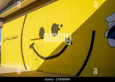 A vibrant yellow wall adorned with an oversized smiley face mural casts a playful shadow at O'village mall at casablanca , Morocco Stock Photo