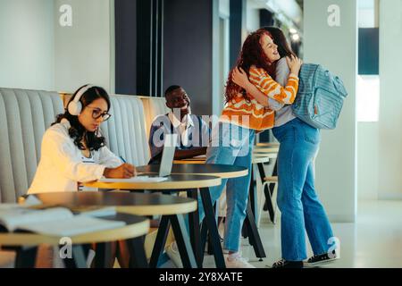 University friends happily reunite with a friendly greeting in a bright, modern study area, fostering academic and social connections among students. Stock Photo