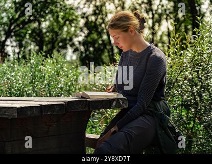 Beautiful woman sits quietly at a wooden table, engaged in a book while surrounded by greenery, enjoying reading in a serene moment in summer nature Stock Photo