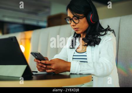 Young student texting on smartphone while studying in a cafe, wearing headphones and using a laptop. A scene of concentration and modern connectivity. Stock Photo