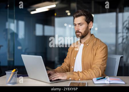 Serious man at work with laptop inside office, thinking businessman typing on keyboard, preparing electronic report, mature worker programmer, coding new software at workplace. Stock Photo