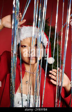 A captivating Asian woman radiates holiday spirit, peeking through colorful decorations. Stock Photo