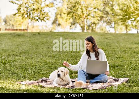 A young woman sits on a blanket in a park, engaged with her laptop while her dog relaxes beside her. Stock Photo