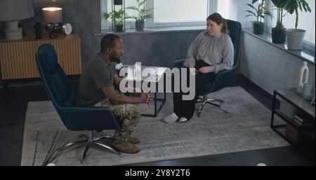 High angle of African American military man with PTSD sitting in chair, talking with psychologist during therapy. Female therapist listens patient on counseling session. Post traumatic stress disorder Stock Photo