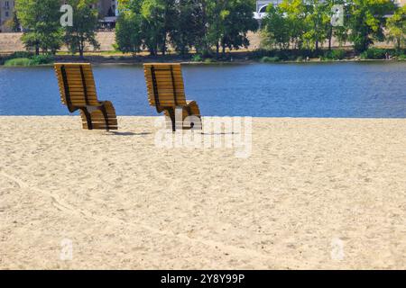 Two wooden sun loungers on a sandy beach. Blue river. Trees on the other side of the river. Stock Photo