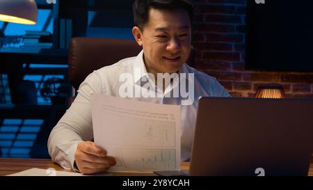 Middle-aged happy smiling businessman working with laptop and documents at dark night office Asian Korean man financial analyst accounting business Stock Photo