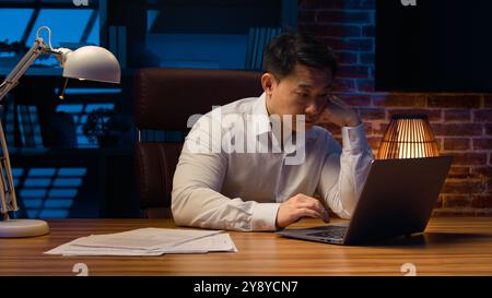 Exhausted fatigued tired Asian man manager office worker at night evening overworked deadline job project working with boring laptop startup sleepy Stock Photo