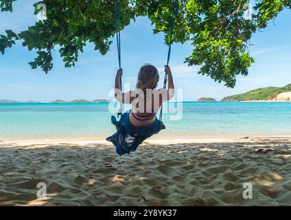 A woman swings on a tropical beach under lush trees, enjoying the serene ocean view on a sunny day Stock Photo
