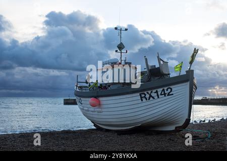 A traditional fishing boat rests on the pebbled shore in Hastings as stormy clouds gather above the tranquil sea. Stock Photo