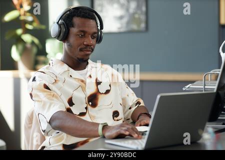 Portrait of trendy African American man using laptop in office and wearing big headphones typing at keyboard and focusing on work, copy space  Stock Photo