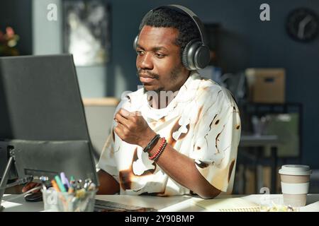 Portrait of adult African American man using computer and wearing big headphones while working at office workplace in sunlight Stock Photo