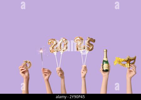 Female hands with figure 2025 made of foil balloons, sparkler, bottle of champagne, Christmas tree decor and carnival mask on lilac background Stock Photo