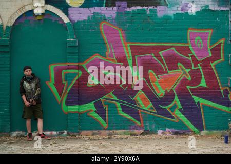 Young creative male graffiti artist standing by green wall of brick building with freshly created artwork painted with various colors Stock Photo