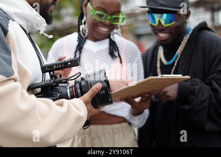 Filming crew holding camera and reviewing script on clipboard during an outdoor film shoot. Team members focused on scene planning without direct eye contact with camera Stock Photo