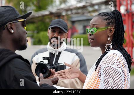 Three people engaging in conversation outdoors with two individuals facing each other and one person holding a camera. One person wears bright green glasses Stock Photo