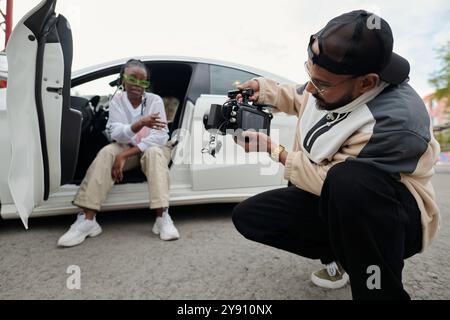 Man holding camera recording woman sitting in car while adjusting settings and angles wearing green glasses and casual outfit capturing outdoor scene without flash Stock Photo