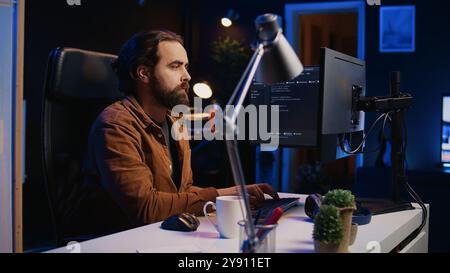 Developer overthinking solutions while doing programming during remote job shift in home office. IT admin working on computer in apartment late at night, thinking how to fix bugs, camera B Stock Photo
