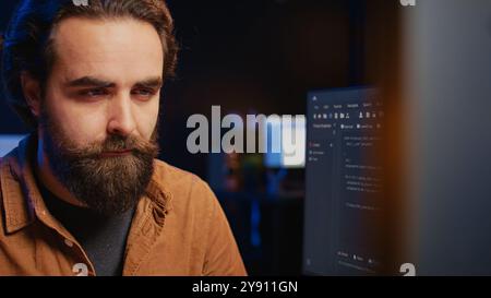 Focused software developer in home office doing coding on computer at desk, close up. IT specialist paying close attention to screen, writing lines of code at workstation, camera B Stock Photo