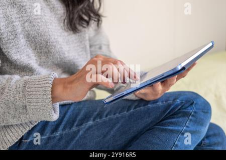 A woman sits casually on a couch, using a digital tablet, her hand tapping the screen, dressed in a cozy sweater and jeans, illustrating relaxed techn Stock Photo