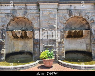A stone wall with two arched alcoves, each containing a small fountain. A potted plant with flowers is placed in front of the wall on a brick surface. Stock Photo