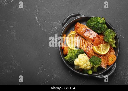 Healthy baked fish salmon steaks, broccoli, cauliflower, carrot in cast iron casserole bowl black dark stone background. Cooking a delicious low carb Stock Photo