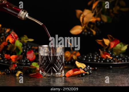 Black chokeberry juice and fresh berries on a black background. Juice or liquor is poured from a bottle into a glass. Stock Photo