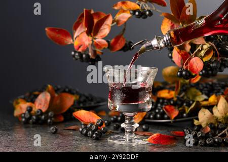 Black chokeberry drink is poured from a bottle into a glass. Sweet drink and fresh berries on a black stone table. Stock Photo