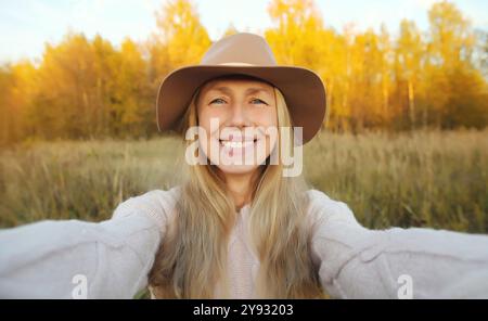 Close up portrait of happy smiling mature woman taking selfie with smartphone, stylish female middle aged model in hat outdoors, warm sunny park, fall Stock Photo