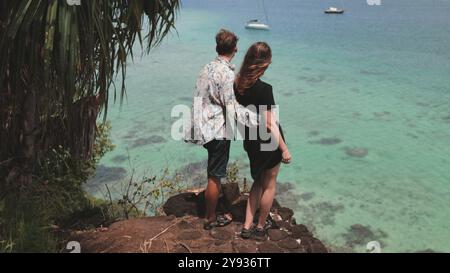 Young couple is embracing on a cliff edge while looking out over a beautiful tropical ocean with sailboats in the distance Stock Photo