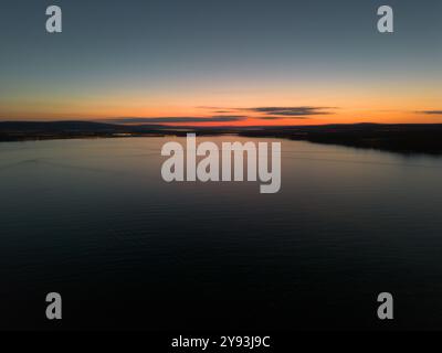 A serene aerial sunset over a calm lake, with subtle reflections on the water and a gradient sky transitioning from deep blue to warm orange hues. Stock Photo