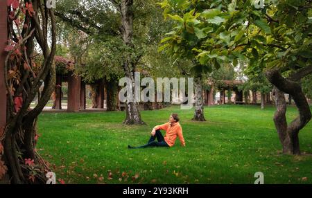 Relaxed man sitting alone on the lush grass in a scenic green park, framed by gnarly trunks and plants Stock Photo