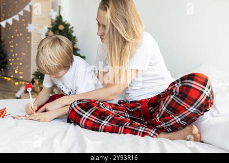 Mother and son in pajamas sitting on the bed write a letter to Santa and dream about gifts. Merry Christmas and Happy Holidays. concept of preparing f Stock Photo