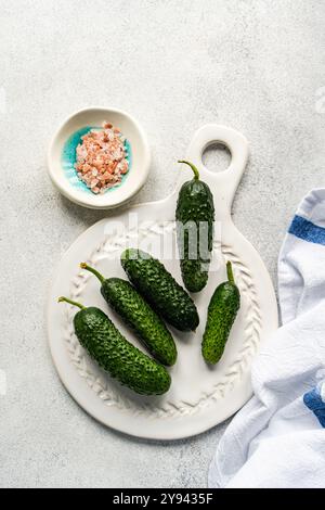 Top view of fresh cucumbers alongside a bowl of pink sea salt, arranged on a white textured serving board. Near the components, a light-colored kitche Stock Photo