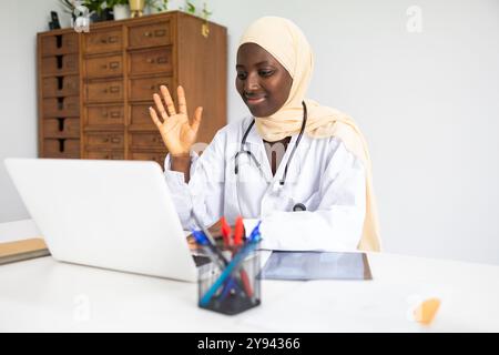 A professional black female doctor wearing a hijab smiles engagingly during a video conversation, her laptop open in front of her in a well-organized Stock Photo