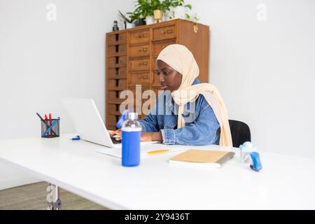A professional black female doctor wearing a hijab is focused on her work at a laptop in a modern office environment Stock Photo