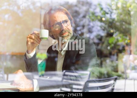 Middle-aged businessman sipping coffee at an outdoor cafe, looking away contemplatively, with a laptop visible on the table. Stock Photo