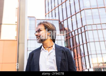 A mature businessman in a stylish black suit looks away thoughtfully, set against a backdrop of modern office buildings. Stock Photo
