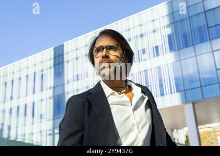 Middle-aged businessman in a suit standing in front of a modern glass building, looking away thoughtfully. Stock Photo