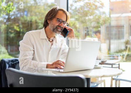 Middle-aged businessman multitasking in a bright cafe, engaging on a laptop while conversing over a mobile phone. He's focused and professional. Stock Photo