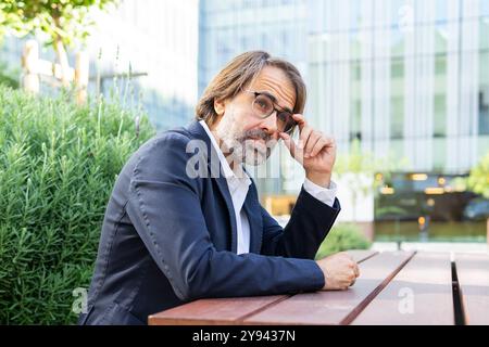 Middle-aged businessman adjusts his glasses while sitting outdoors in a city, with modern buildings in the background. Stock Photo