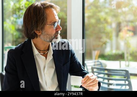 A middle-aged man looks thoughtfully through a large window in a bright city office while holding a white coffee cup, dressed in a smart black suit an Stock Photo
