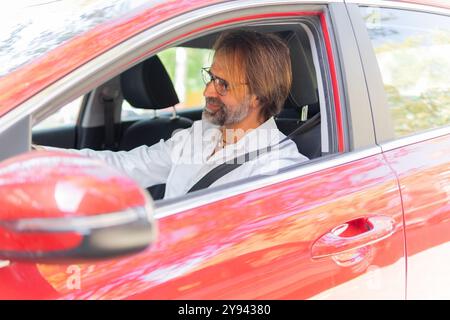 Side view of a businessman with a beard and glasses, wearing a white shirt, is seen using a laptop while sitting in the driver's seat of a red car. Stock Photo