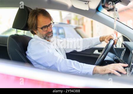 A mature businessman in a white shirt adjusts the controls in his car while looking down, focusing on his driving tasks. Stock Photo