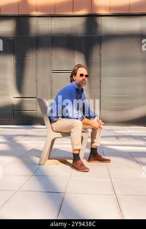 A middle-aged businessman in a blue shirt and beige pants sits relaxed on a modern bench in an urban setting, looking away thoughtfully. Stock Photo