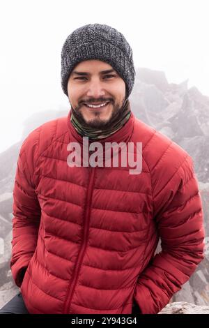A young man in a red insulated jacket smiles at the camera while on a cloudy mountain trail. He wears a gray knitted hat and a scarf, enhancing the co Stock Photo