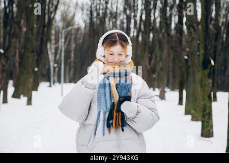 A young teenage girl braves the cold winter weather, cozily dressed in a thick scarf, jacket, and mittens, while listening to music through white head Stock Photo