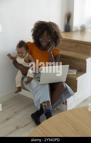 An African mother smiles while cradling her baby girl on her lap and working on a laptop, exemplifying modern parenting in a cozy home setting Stock Photo