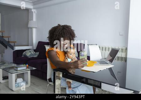 African mother sits at a modern home dining table, smiling while interacting with her baby girl using a smartphone Stock Photo